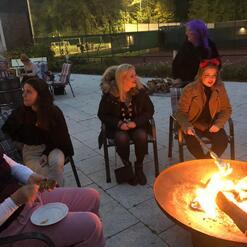 Gruppe von Frauen sitzt um ein Feuer, einige r&ouml;sten Snacks, entspannte Atmosph&auml;re im Au&szlig;enbereich.