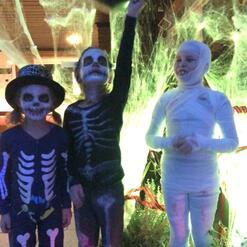 Drei Kinder in Halloween-Kost&uuml;men: zwei als Skelette, eines als Mumie, vor gruseliger Dekoration.