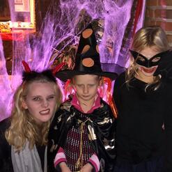 Drei Kinder in Halloween-Kost&uuml;men stehen vor einer dekorierten Wand mit Spinnennetzen und gruseliger Beleuchtung.
