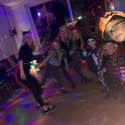 Gruppe von Personen in Kost&uuml;men tanzt in einem festlich beleuchteten Raum, Halloween-Dekoration im Hintergrund.