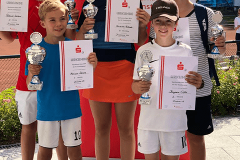 Gruppe von f&uuml;nf jungen Sportlern mit Pokalen und Urkunden vor einem roten Banner. Alle l&auml;cheln auf einem Tennisplatz.