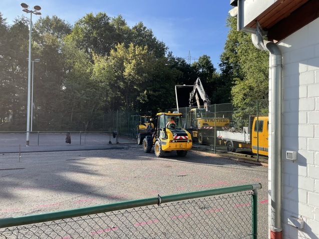 Bauarbeiten auf einem Platz mit Gelben Maschinen und einem gro&szlig;en Bagger hinter einem Zaun, umgeben von B&auml;umen.