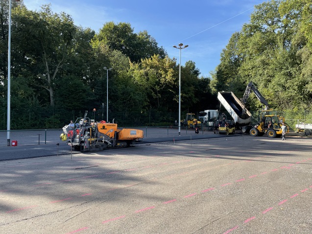 Blick auf eine Baustelle mit verschiedenen Stra&szlig;enbaumaschinen und B&auml;umen im Hintergrund.