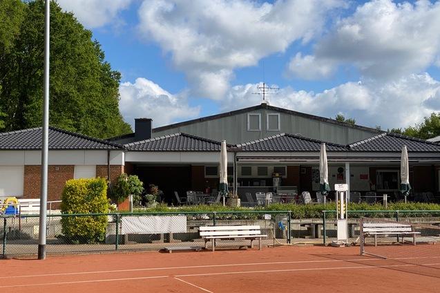 Tennisplatz mit einem Vereinsgeb&auml;ude im Hintergrund und Sonnenschirmen auf der Terrasse, umgeben von B&auml;umen und Wolken.