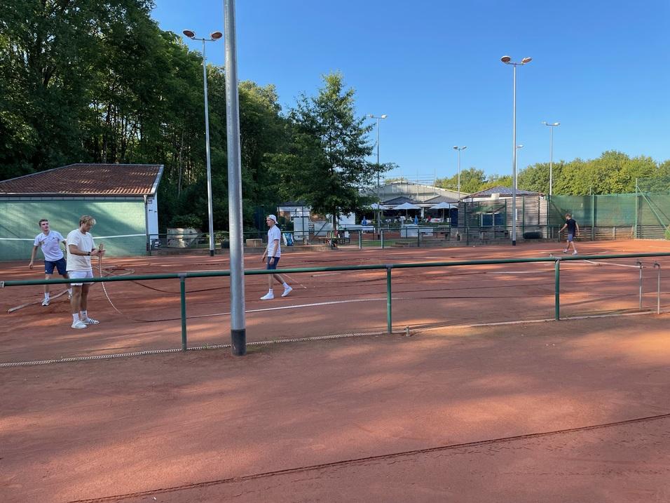 Zwei Personen spielen Tennis auf einem Sandplatz, während andere im Hintergrund aktiv sind und ein Vereinsgebäude sichtbar ist.