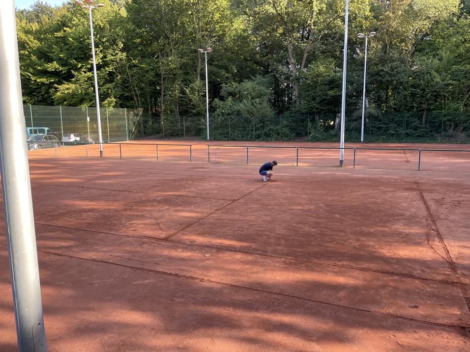 Ein Kind kniet auf einem Tennisplatz aus roter Erde, umgeben von grünen Bäumen und Sichtschutz.