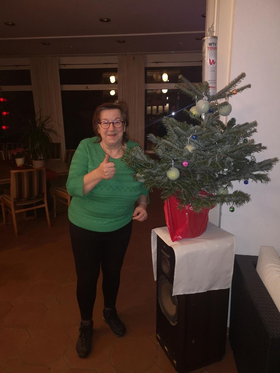 Frau in Grün zeigt Daumen hoch neben einem kleinen Weihnachtbaum in einem Raum mit Stühlen und Beleuchtung.