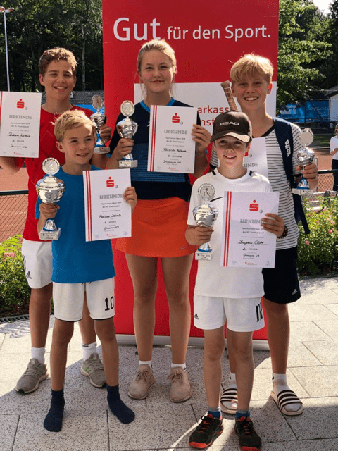 Gruppe von fünf jungen Sportlern mit Pokalen und Urkunden vor einem roten Banner. Alle lächeln auf einem Tennisplatz.