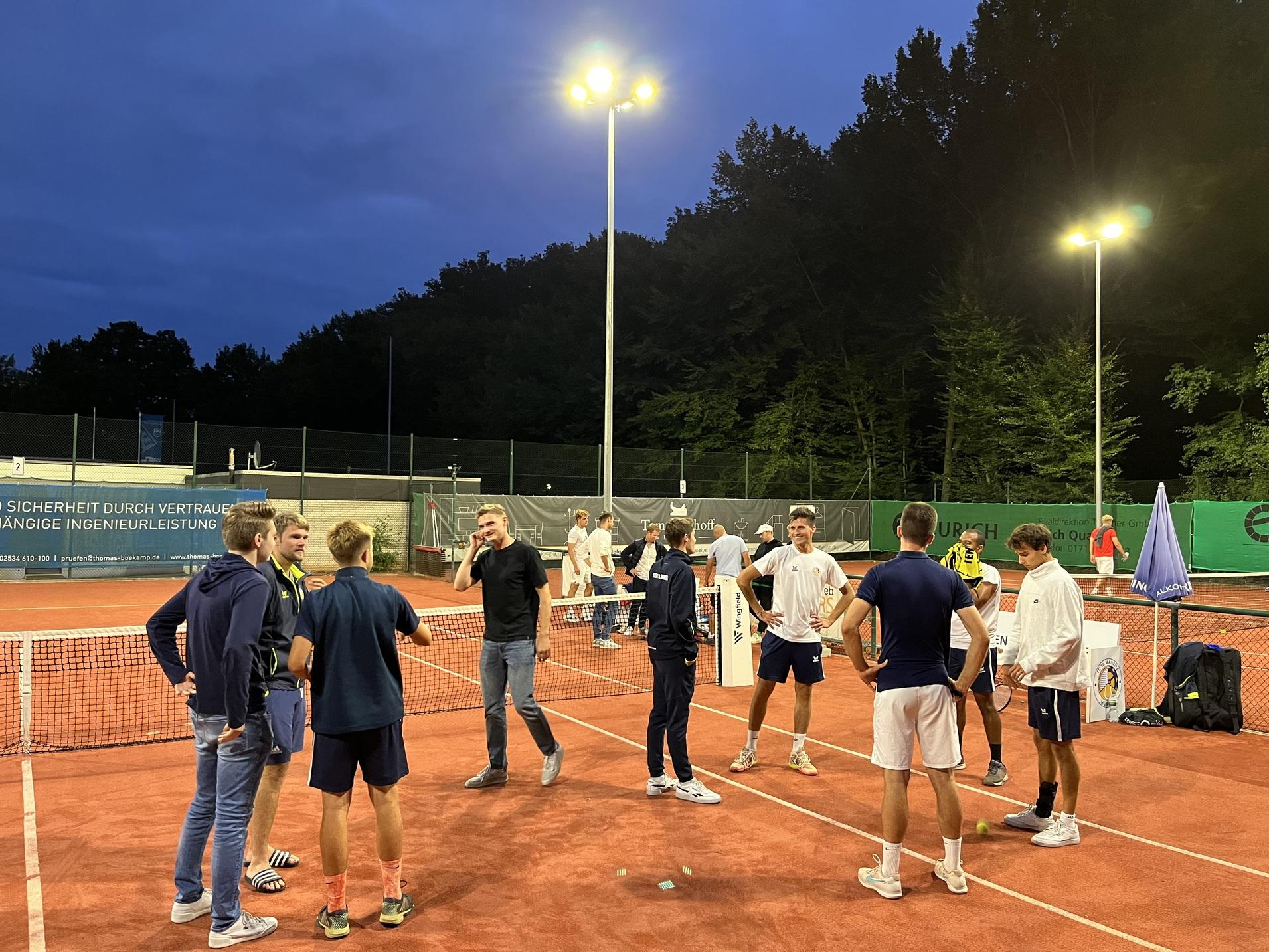 Gruppe junger Männer unterhält sich auf einem beleuchteten Tennisplatz bei Abenddämmerung.