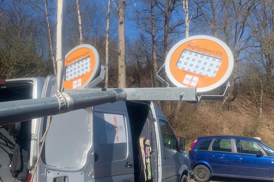 Transporter mit offener Hintert&uuml;r und montierten LED-Scheinwerfern unter blauem Himmel, umgeben von B&auml;umen.