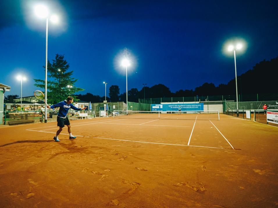 Tennisspieler schl&auml;gt in der D&auml;mmerung auf einem beleuchteten Sandplatz.