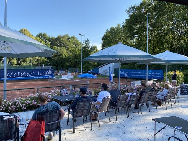 Blick auf einen Tennisplatz mit Zuschauern in St&uuml;hlen unter Schirmen, umgeben von Blumenbeeten und B&auml;umen.