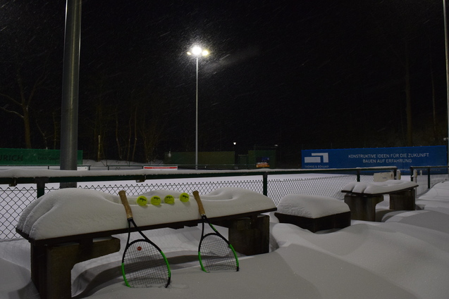 Zwei Tennisschl&auml;ger und gelbe B&auml;lle liegen auf einer bei Nacht schneebedeckten Bank an einem Tennisplatz.
