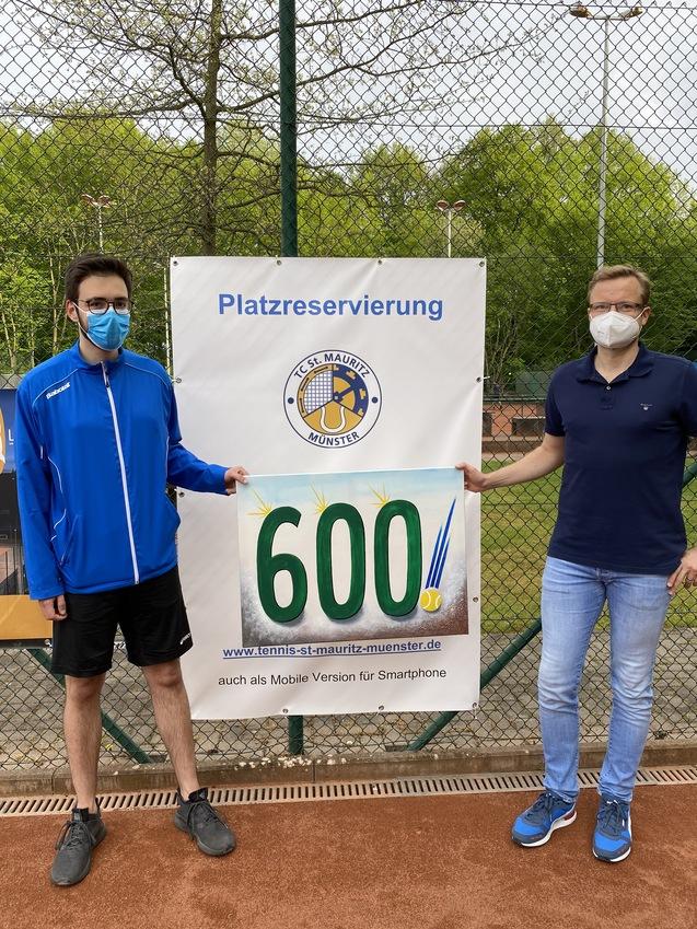 Zwei M&auml;nner in Masken stehen vor einem Schild mit der Aufschrift "Platzreservierung" und der Zahl "600" auf einem Tennisplatz.