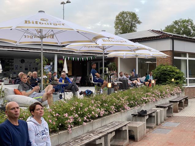 Gruppe von Menschen sitzt unter Schirmen an einem Caf&eacute; im Freien, umgeben von Blumen und einer entspannenden Atmosph&auml;re.