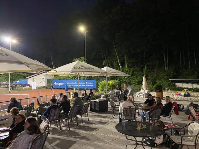Personen sitzen abends an Tischen unter Sonnenschirmen neben einem Tennisplatz, umgeben von B&auml;umen und Beleuchtung.