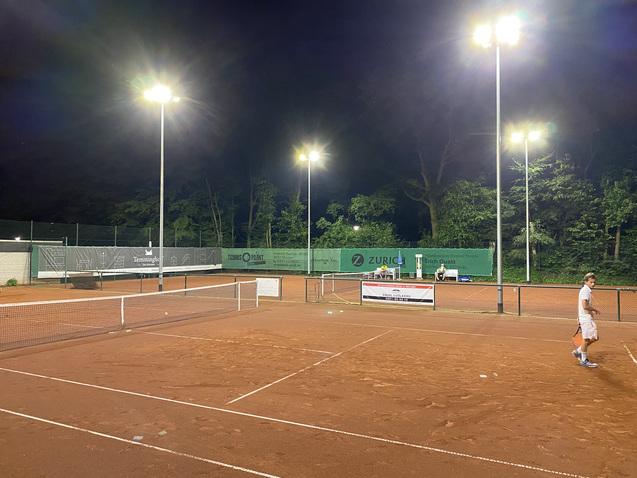 Tennisplatz bei Nacht mit Beleuchtung, zwei Spielern und Werbebannern im Hintergrund. Rote Sandoberfl&auml;che und gr&uuml;ne Umgebung.