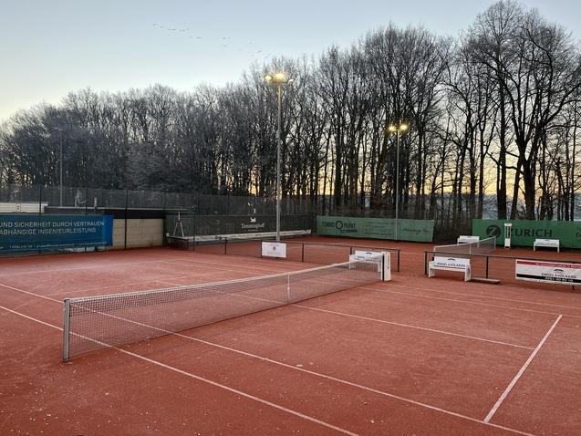 Tennisplatz mit roten Spielfl&auml;chen, Netz in der Mitte und B&auml;umen im Hintergrund bei Sonnenaufgang.