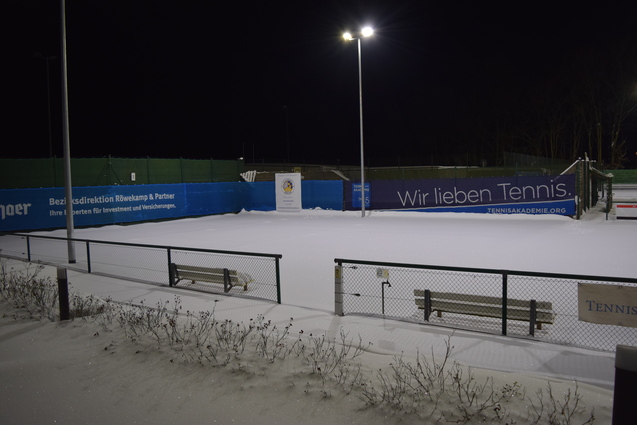 Winterliche Tennisanlage mit schneebedecktem Platz und beleuchteten Netzen bei Nacht. Sicht auf B&auml;nke und Umz&auml;unung.