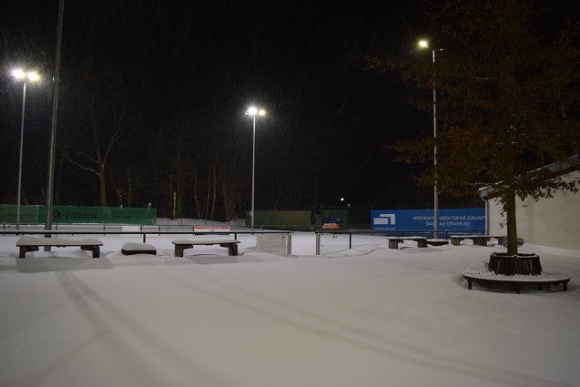 Winterliche Tennispl&auml;tze, bedeckt mit frischem Schnee, unter flackernden Lichtern bei Nacht.