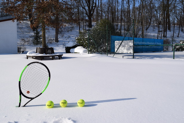 Tennisrakete und drei Tennisb&auml;lle stehen im Schnee vor einem verschneiten Tennisplatz und einem Geb&auml;ude.
