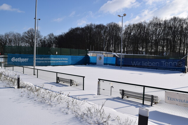 Winterliche Tennisplatzansicht mit Schnee bedeckt, umgeben von B&auml;umen und Werbebannern, keine Spieler sichtbar.