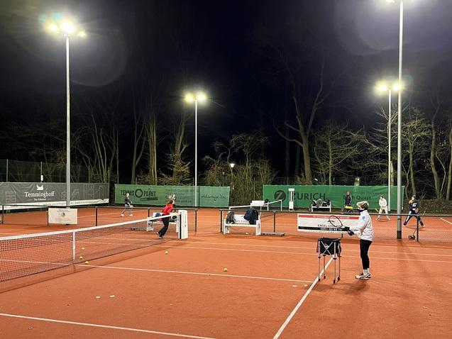 Tennisplatz mit beleuchteten Spielfeldern, mehreren Spielern und Ballmaschinen unter Nachtbedingungen.