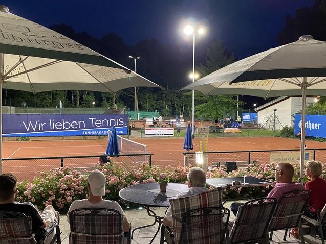 Blick auf einen Tennisplatz bei Nacht, mit Zuschauern unter Sonnenschirmen und Blumenbeeten im Vordergrund.