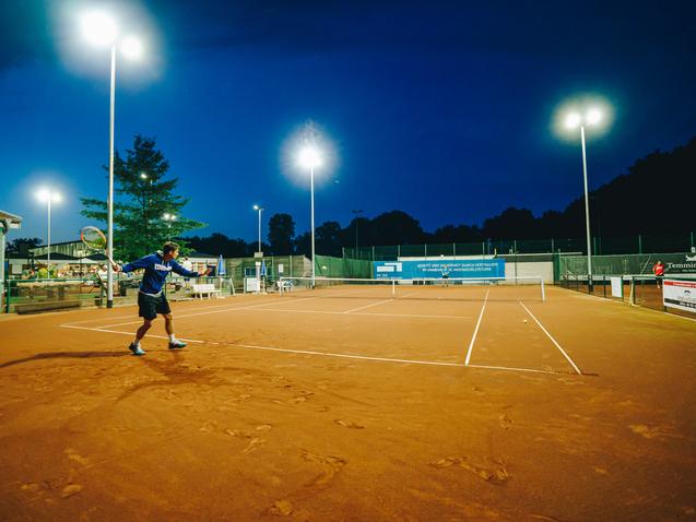 Tennisspieler schl&auml;gt in der D&auml;mmerung auf einem beleuchteten Sandplatz.