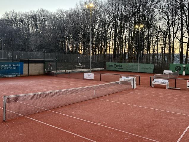 Tennisplatz mit roten Spielflächen, Netz in der Mitte und Bäumen im Hintergrund bei Sonnenaufgang.