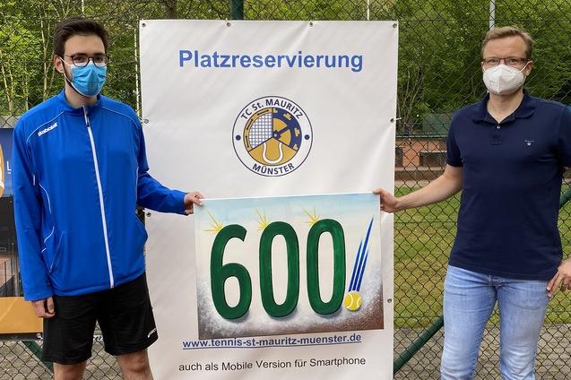 Zwei M&auml;nner in Masken stehen vor einem Schild mit der Aufschrift "Platzreservierung" und der Zahl "600" auf einem Tennisplatz.