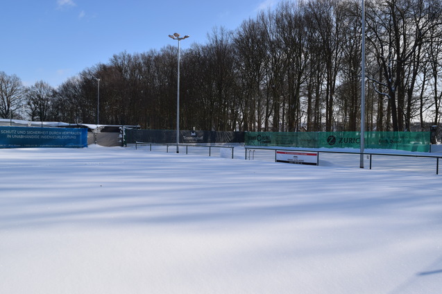Wei&szlig;er Schnee bedeckt eine Sportanlage mit Z&auml;unen und gr&uuml;nen Planen im Hintergrund, umgeben von schneebedeckten B&auml;umen.