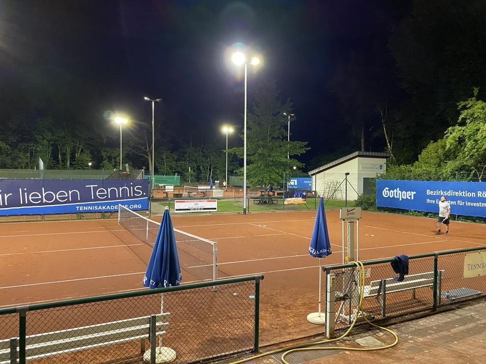 Tennisplatz bei Nacht mit beleuchteten Netzen, einem Spieler und blauen Sonnenschirmen. Werbebanner im Hintergrund.