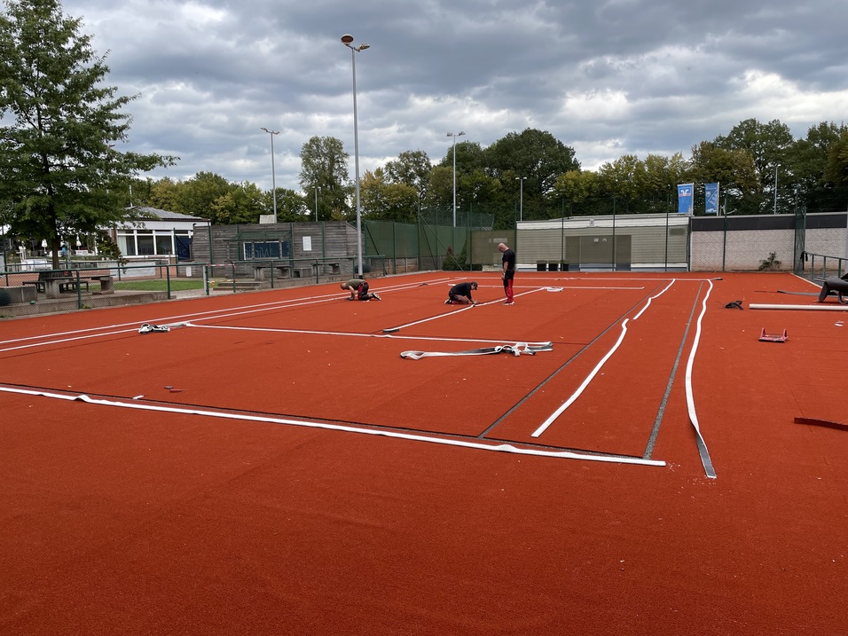 Arbeiter verlegen Linien auf einem roten Tennisplatz unter bew&ouml;lktem Himmel. Im Hintergrund sind B&auml;ume und Geb&auml;ude sichtbar.