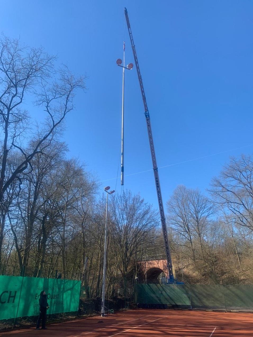 Kran hebt eine hohe Lichtmast auf einem Tennisplatz, umgeben von Bäumen und blauem Himmel.