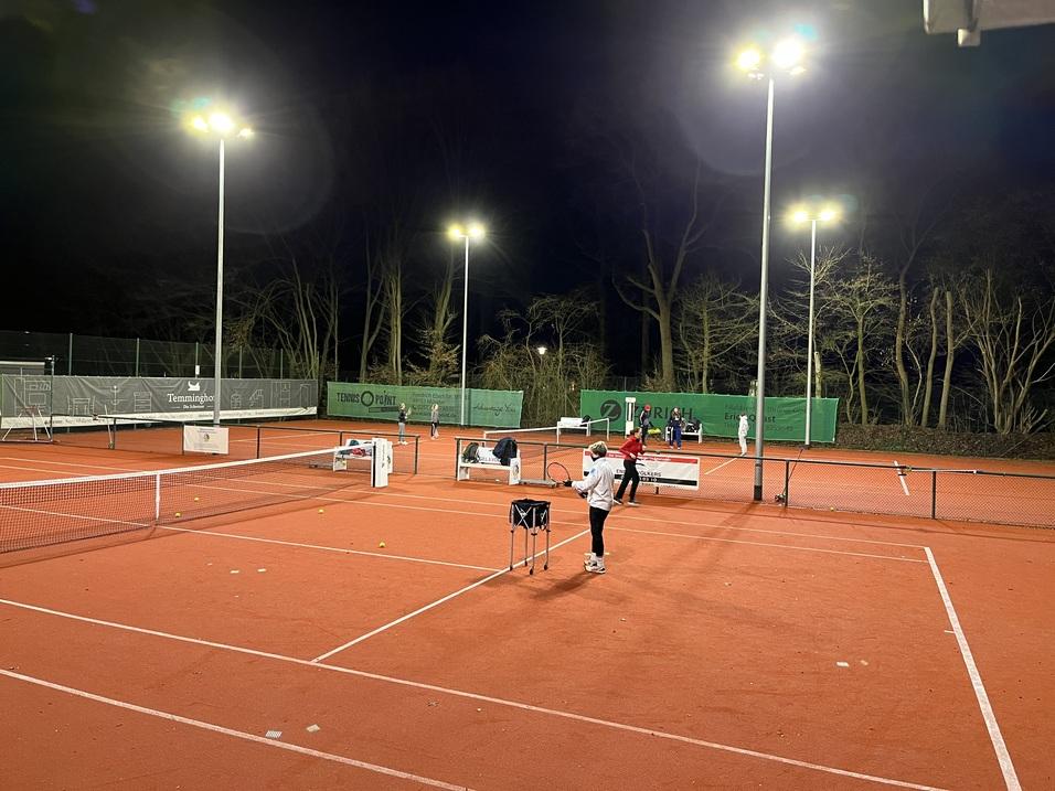 Beleuchteter Tennisplatz in der Nacht mit mehreren Spielern und einem Tisch zum Training.