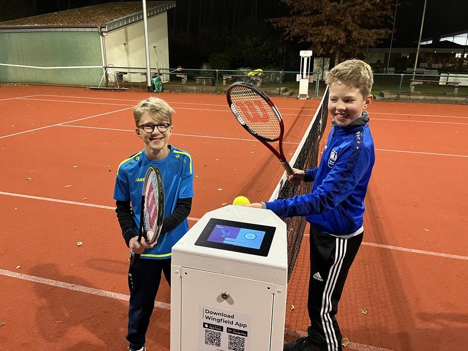 Zwei Kinder halten Tennisschl&auml;ger und stehen an einem Infostand auf einem beleuchteten Tennisplatz.