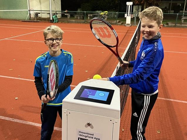 Zwei Kinder halten Tennisschläger und stehen an einem Infostand auf einem beleuchteten Tennisplatz.