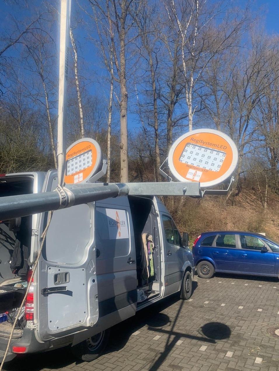 Transporter mit offener Hintertür und montierten LED-Scheinwerfern unter blauem Himmel, umgeben von Bäumen.