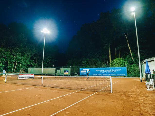 Beleuchteter Tennisplatz mit roten Sand, B&auml;nken und Werbebanner unter einem n&auml;chtlichen Himmel.