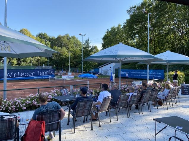 Blick auf einen Tennisplatz mit Zuschauern in St&uuml;hlen unter Schirmen, umgeben von Blumenbeeten und B&auml;umen.