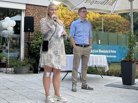 Frau mit Mikrofon spricht bei einer Veranstaltung, Mann im blauen Hemd steht daneben, im Hintergrund eine Terrasse mit Dekoration.
