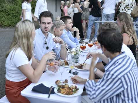 Gesellige Runde von Personen, die an Stehtischen Speisen und Cocktails genie&szlig;en, vor einer ungezwungenen Veranstaltung im Freien.
