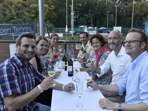 Gruppe von sieben Personen sitzt an einem Tisch, hebt Gl&auml;ser und feiert auf einem Tennisplatz im Freien.