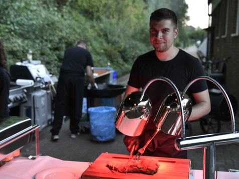 Mann mit Fleischschneider an einem Tisch im Freien, w&auml;hrend ein Grill im Hintergrund raucht.