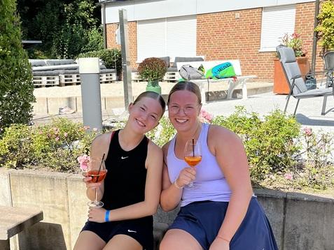 Zwei junge Frauen sitzen unter einem Schirm, halten Getränke und lächeln in einem sonnigen Garten.