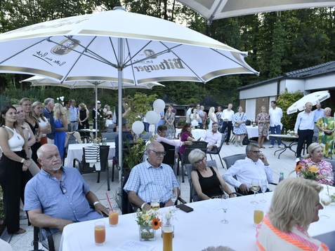 Gruppierung von Menschen in einem Garten, umgeben von St&uuml;hlen und Tischen, unter Sonnenschirmen, bei einem festlichen Anlass.