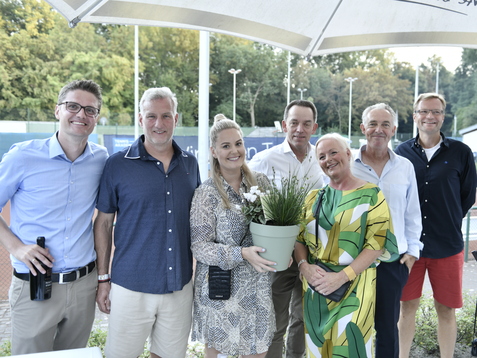 Gruppennahaufnahme von sechs Personen, die zusammen mit einem Blumentopf auf einer Tennisanlage posieren.