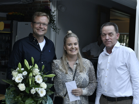 Drei Personen stehen zusammen, eine h&auml;lt einen Blumenstrau&szlig;, die anderen halten eine Karte in einem Innenraum.