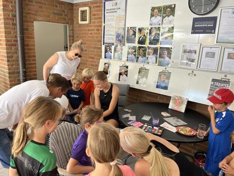 Gruppenszene von Kindern und Erwachsenen, die sich um einen Tisch versammeln, an der Wand Plakate und Bilder.